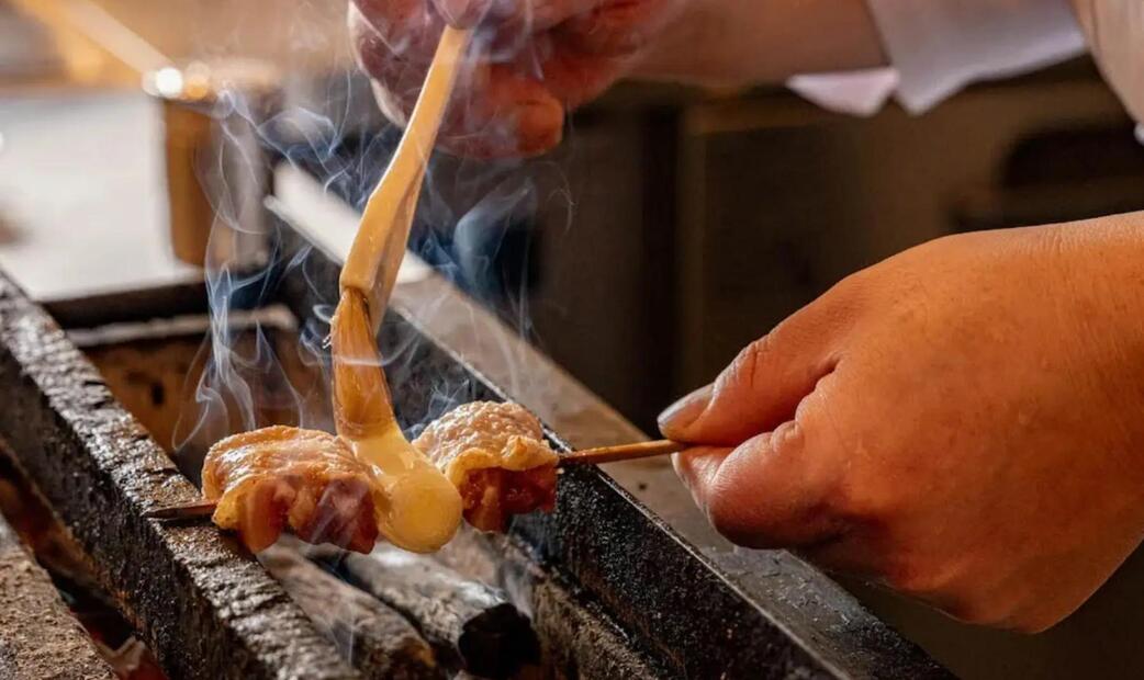 Hands grilling skewered meat over a charcoal fire.