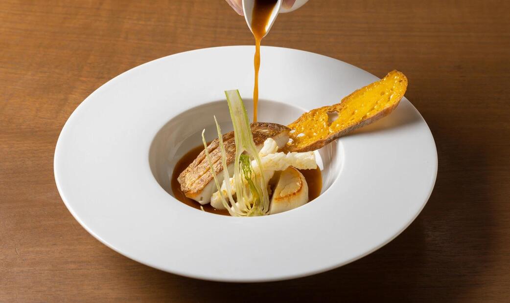 Sauce being poured over fish and vegetables in a white bowl.