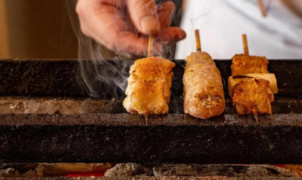 Skewers of grilled meat being cooked over a charcoal grill with a hand turning one of them.